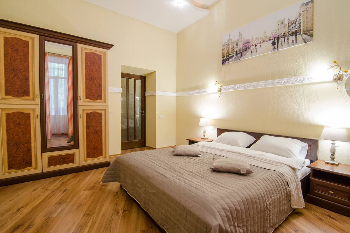 A hotel room with a large bed, wooden wardrobe, and a painting of a cityscape on the wall.