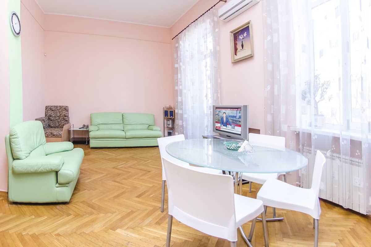 A living room with a green sofa, a glass table with chairs, a television, and a window with sheer curtains.