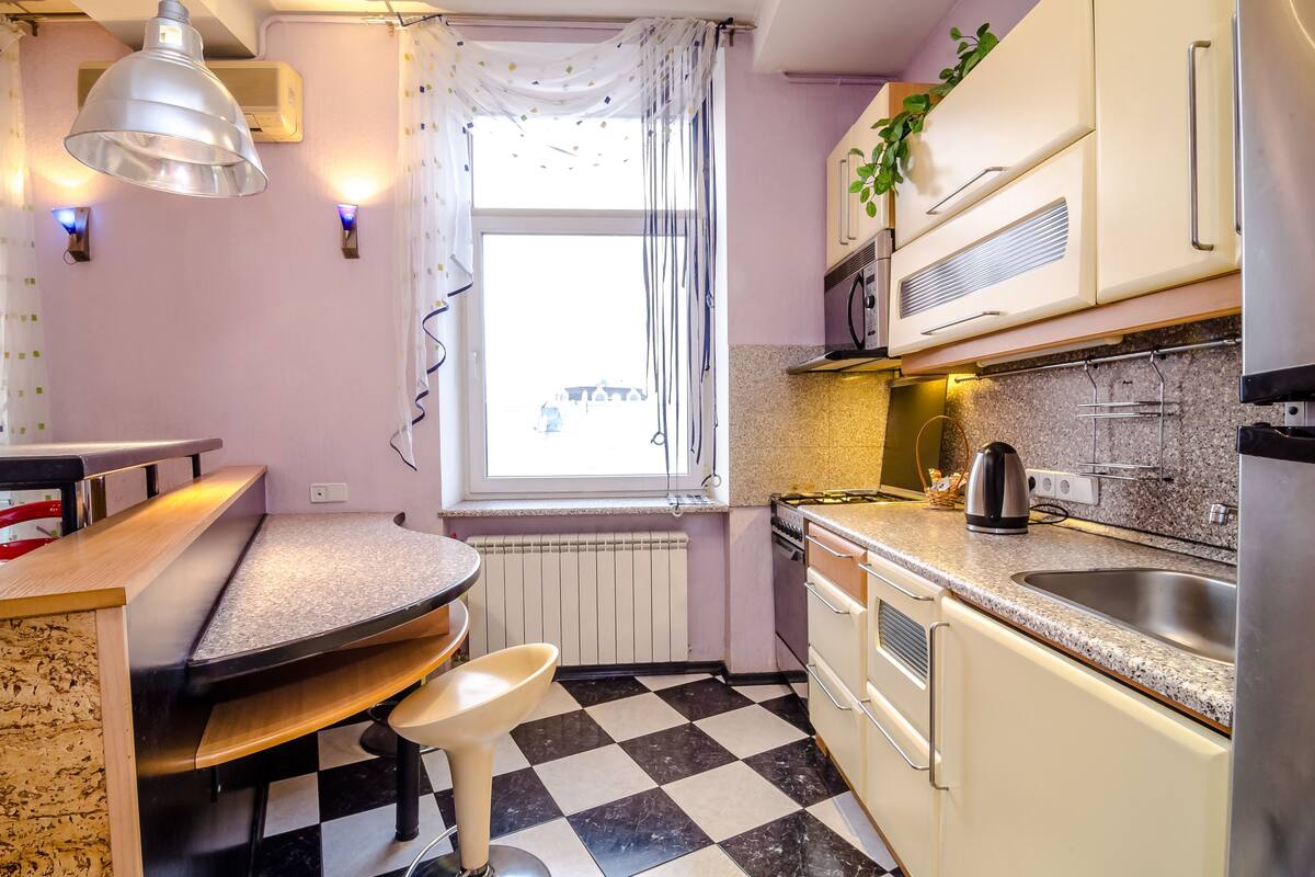 A compact kitchen with a checkered floor, a window with curtains, a kettle, and a sink.