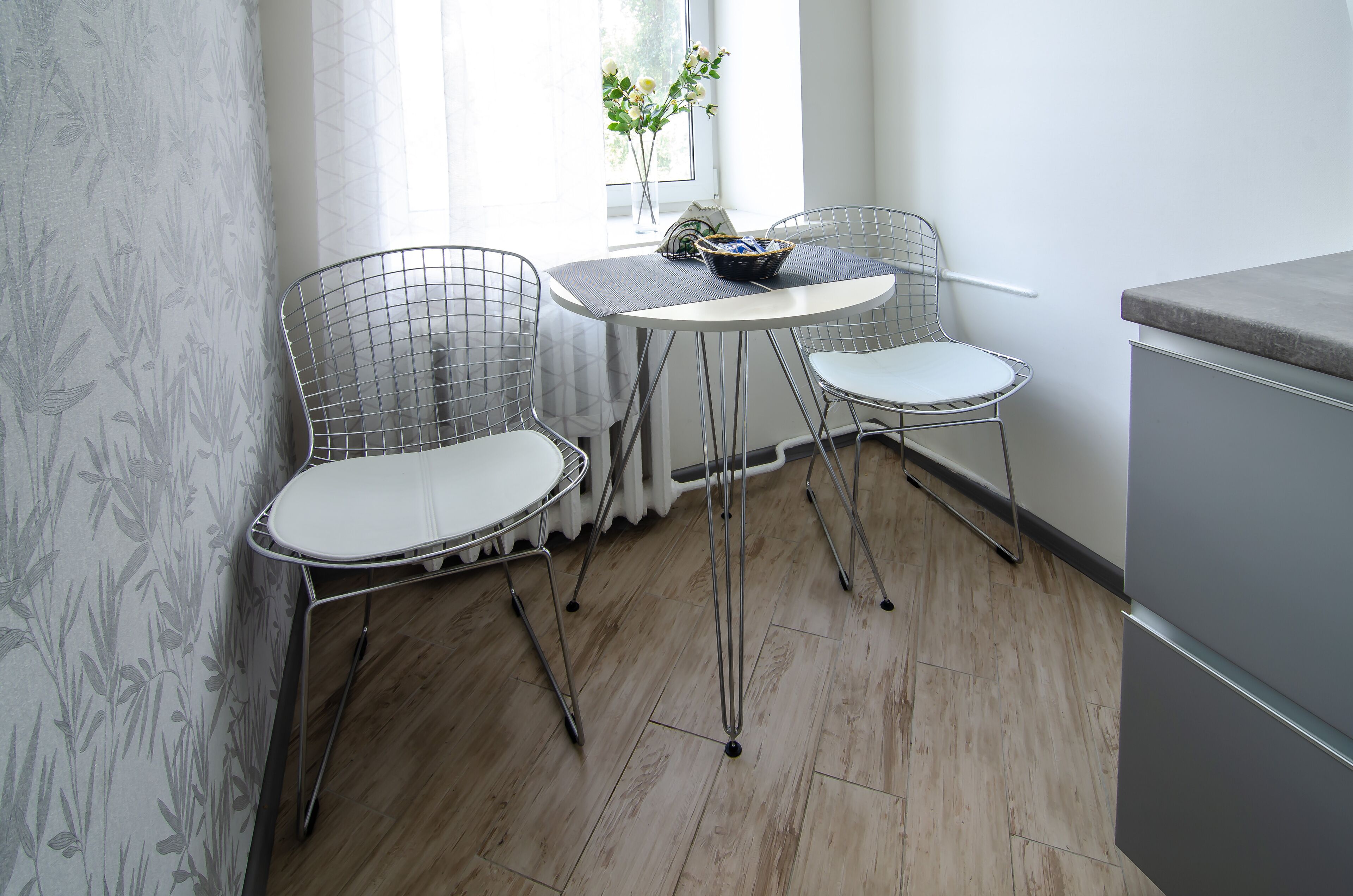 A small dining area with two wire-frame chairs, a round table, and a vase with flowers.