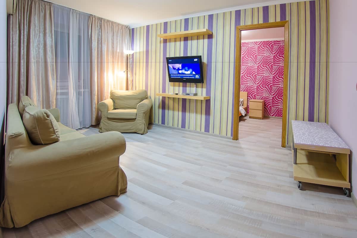 A living room with a beige sofa, a smaller armchair, a flat-screen TV mounted on a wooden shelf, and a patterned wallpaper.