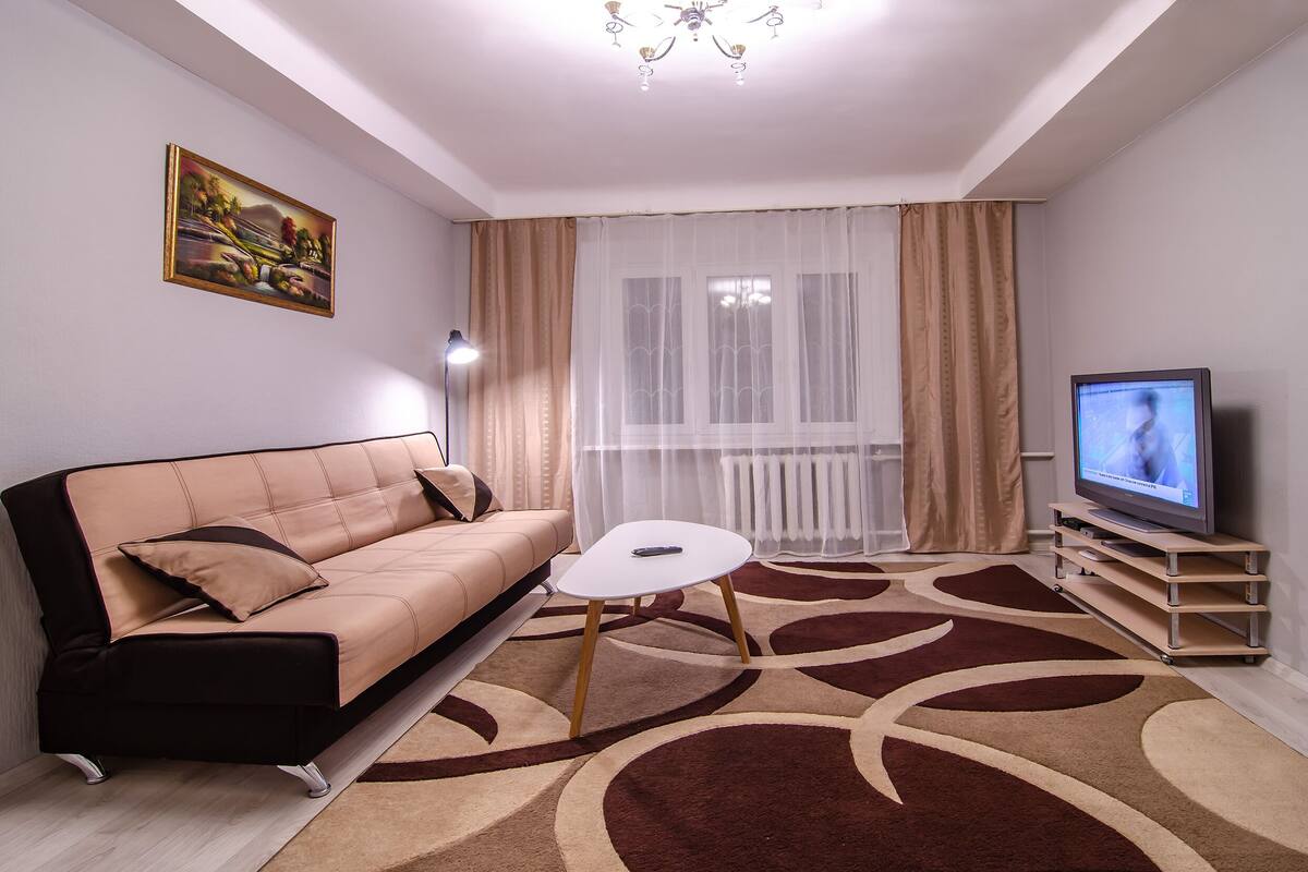 A modern living room with a sofa, a coffee table, a TV on a stand, and a large window with sheer curtains.