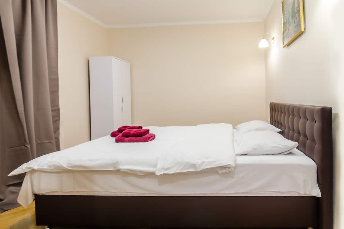 A neatly made bed with white linens and a red towel, a dark brown headboard, a white wardrobe, and a painting on the wall.