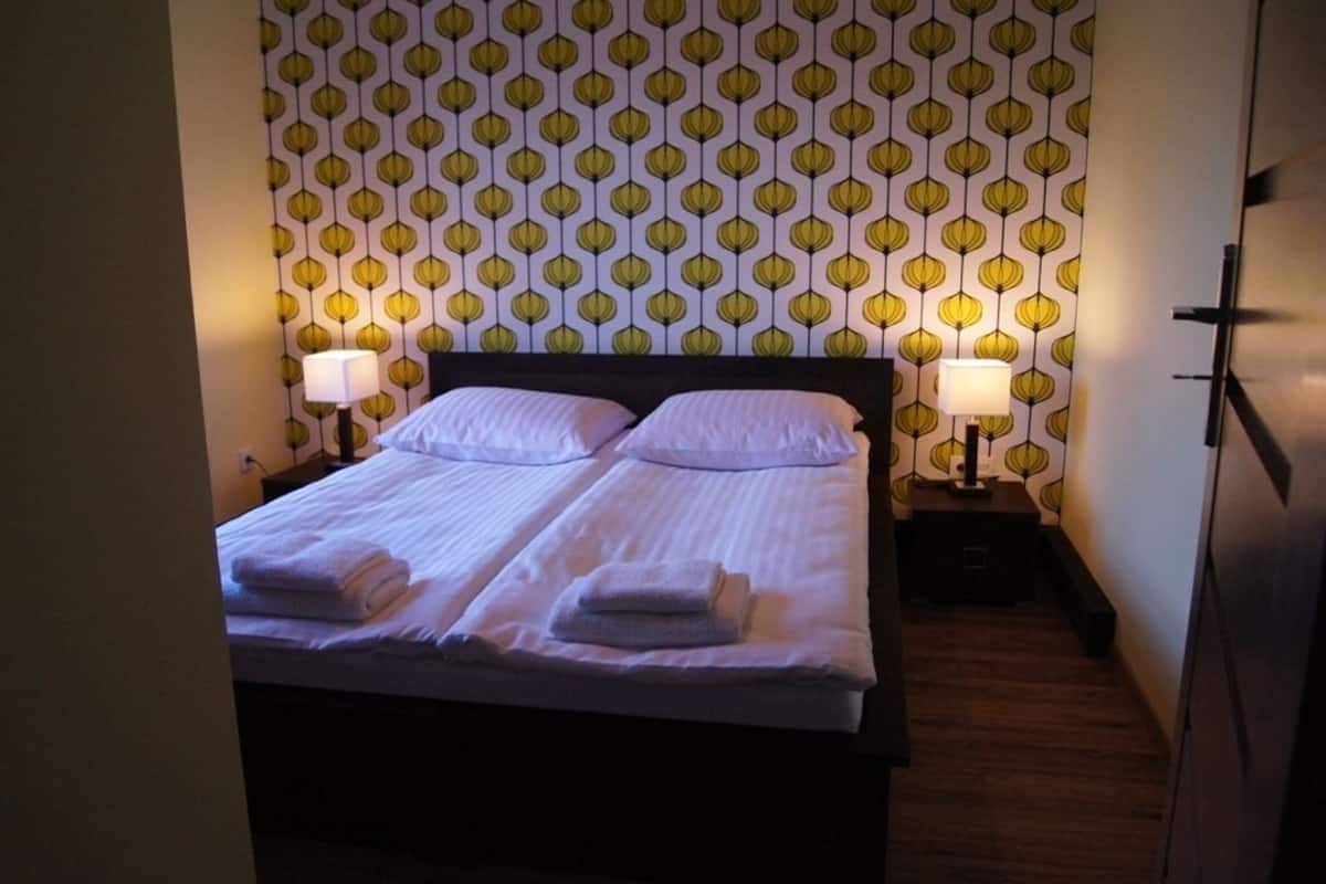 En dobbeltseng med to puder og to sammenfoldede håndklæder, sengeborde med lamper, og tapet med mønster.