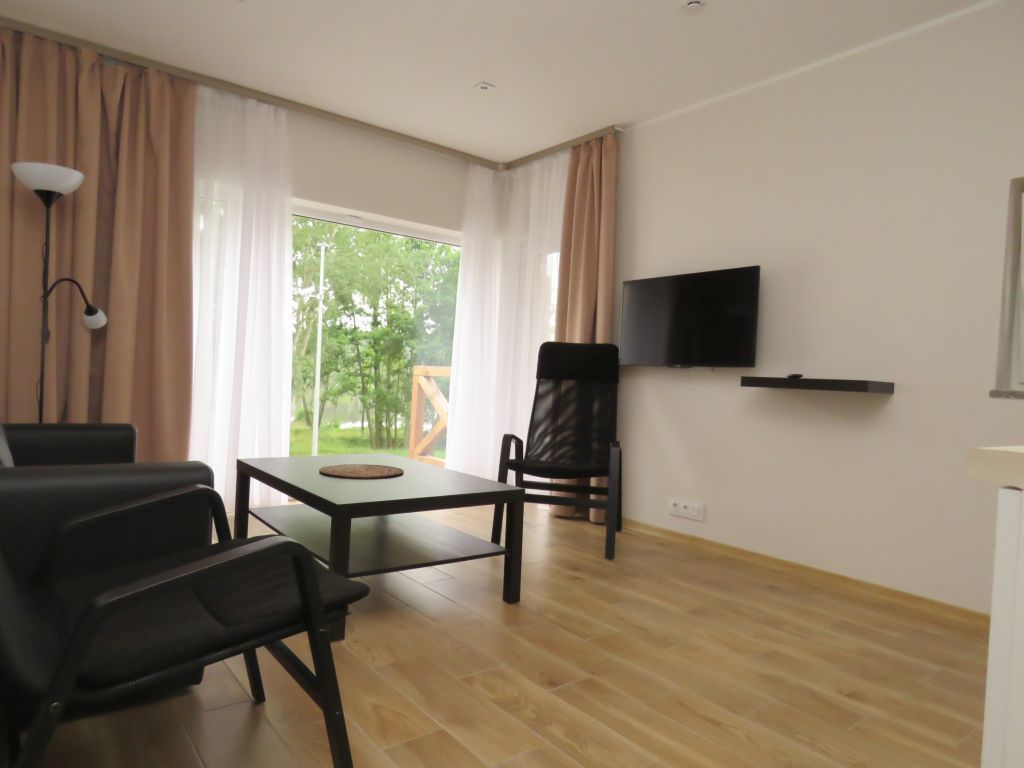 A modern living room with a wooden floor, a dining table with chairs, a flat-screen TV mounted on the wall, and a large window with curtains.