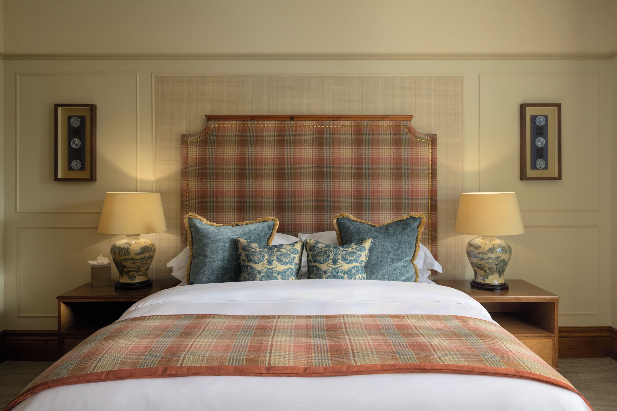 A hotel room with a large bed, two bedside tables with lamps, a headboard with a plaid cover, and framed pictures on the walls.