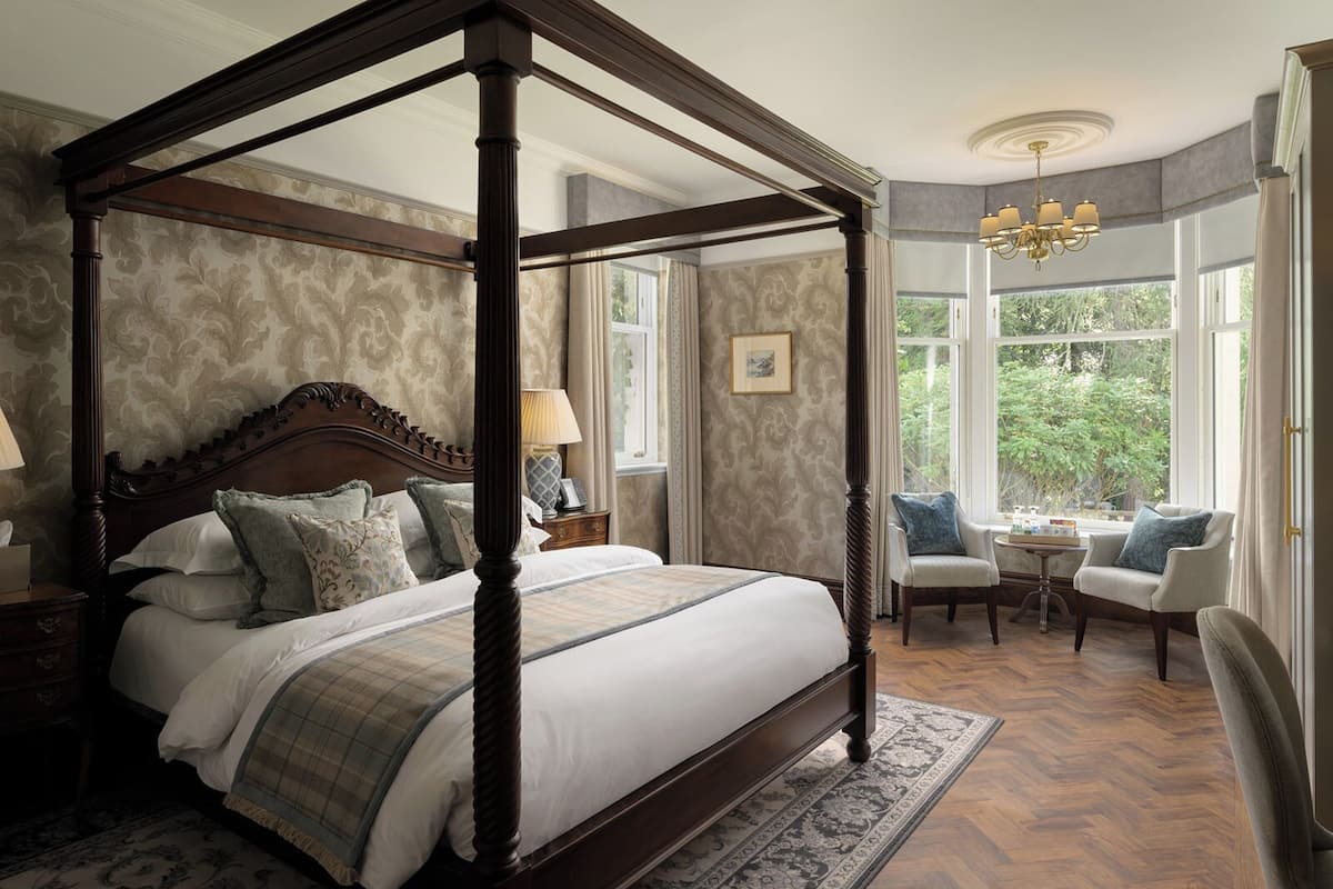 A four-poster bed with a wooden frame, a matching nightstand, a chair, and a table with a lamp.