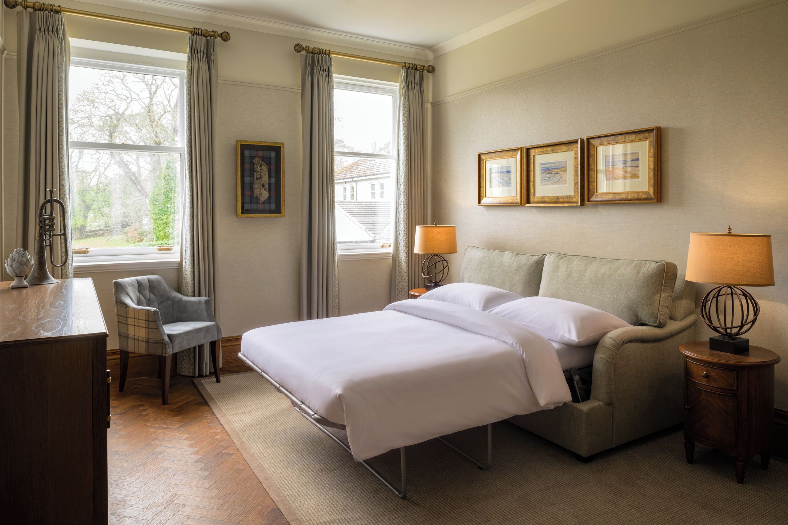 A hotel room with a bed, a chair, a desk, a lamp, and framed pictures on the wall.
