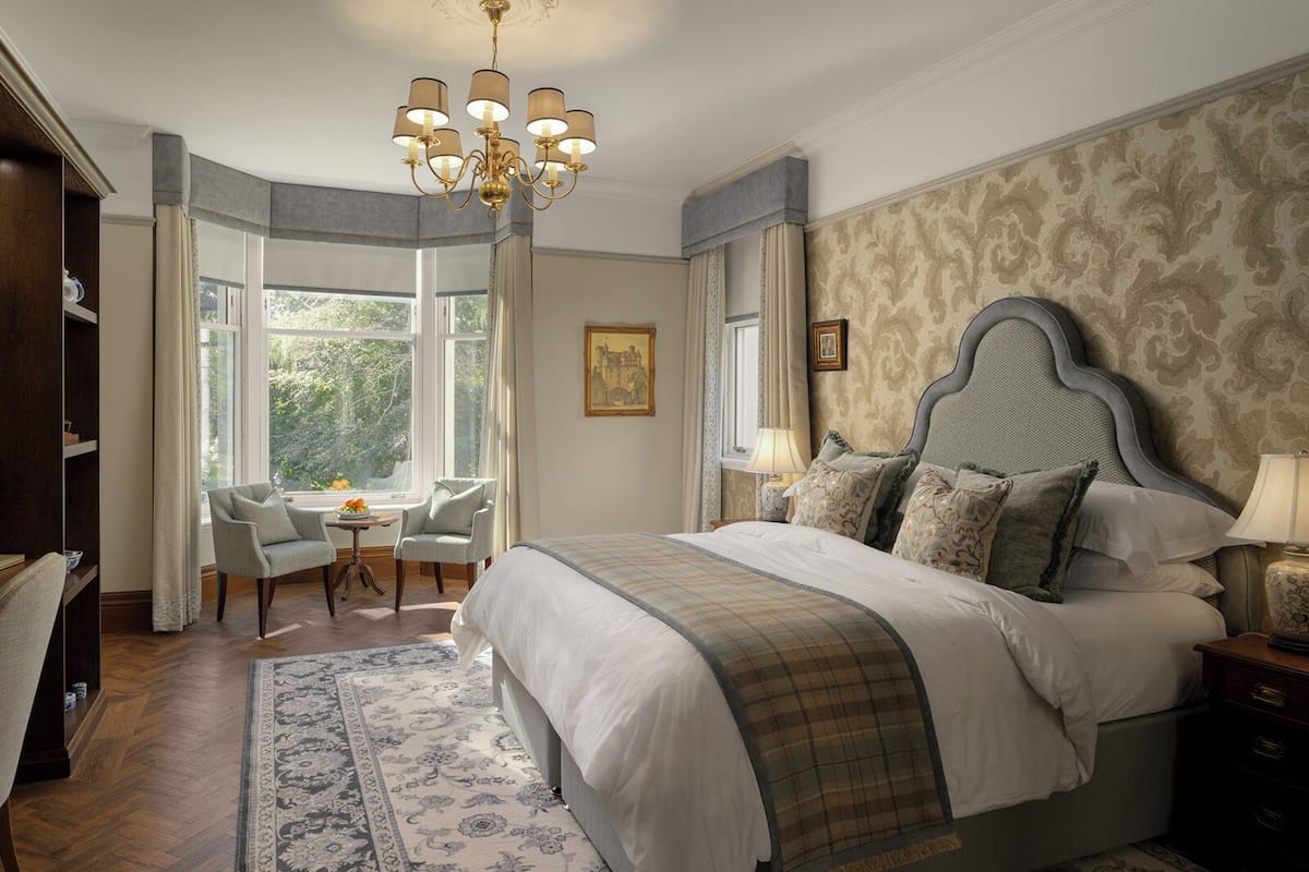 A bedroom with a large bed, a chandelier, a desk with a chair, and a window with curtains.