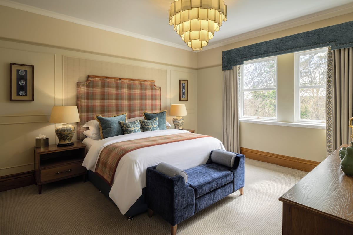 A hotel room with a large bed, two bedside tables, a nightstand, a bench, a chandelier, and a window with curtains.