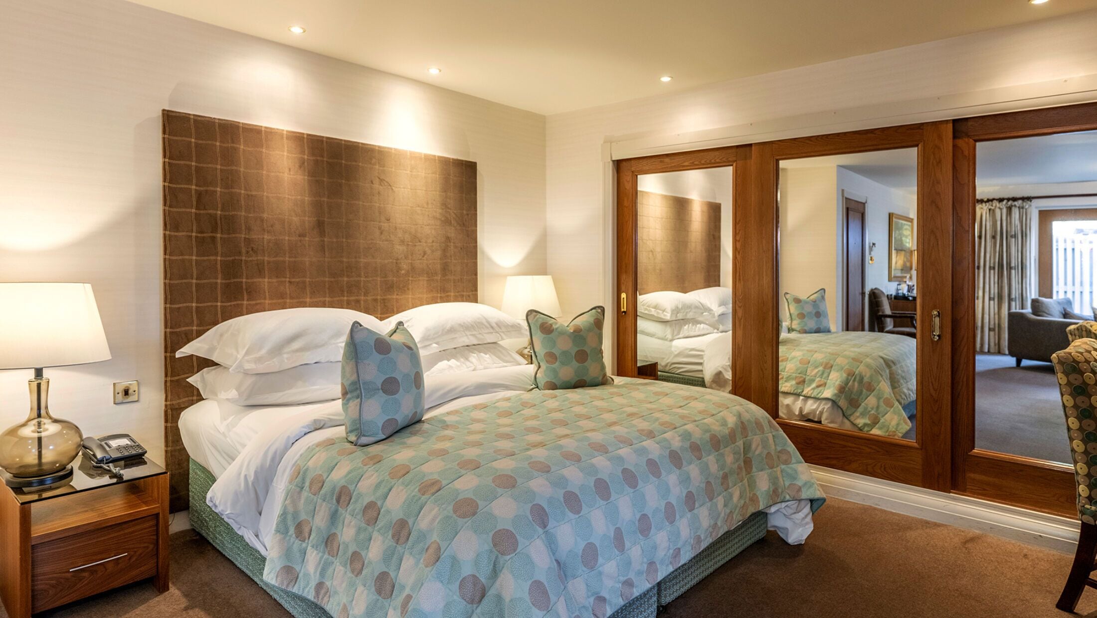 A hotel room with a large bed, bedside lamps, a telephone, a mirror, and a view of the adjacent living area.