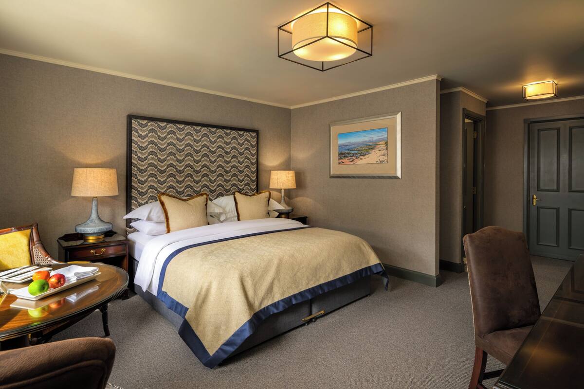 A hotel room with a bed, two bedside tables, a desk with a chair, a lamp, a framed picture on the wall, and a door.