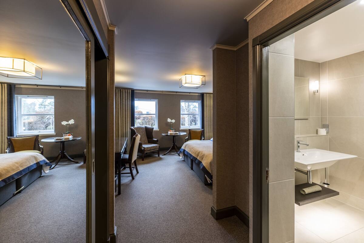 A hotel room with two beds, a desk, a chair, a round table, and a bathroom with a sink and mirror.