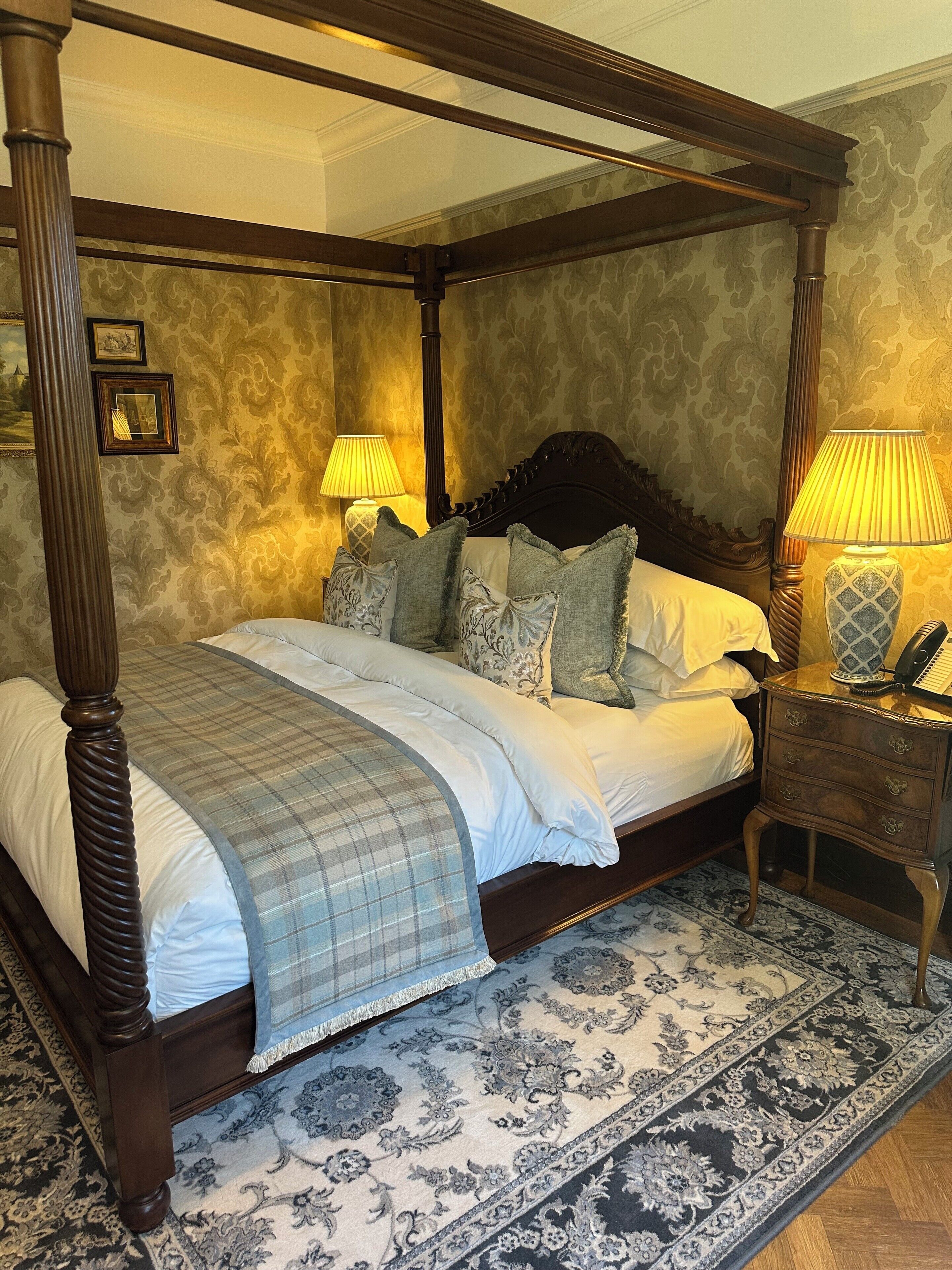 A four-poster bed with a dark wooden frame, a plaid bedspread, and multiple pillows. There are two bedside lamps with lampshades, a small bedside table with a vase, and a patterned rug on the floor.
