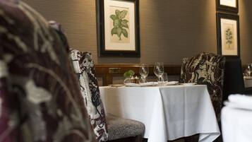 A dining area with a table set for two, framed botanical prints on the wall, and patterned upholstered chairs.