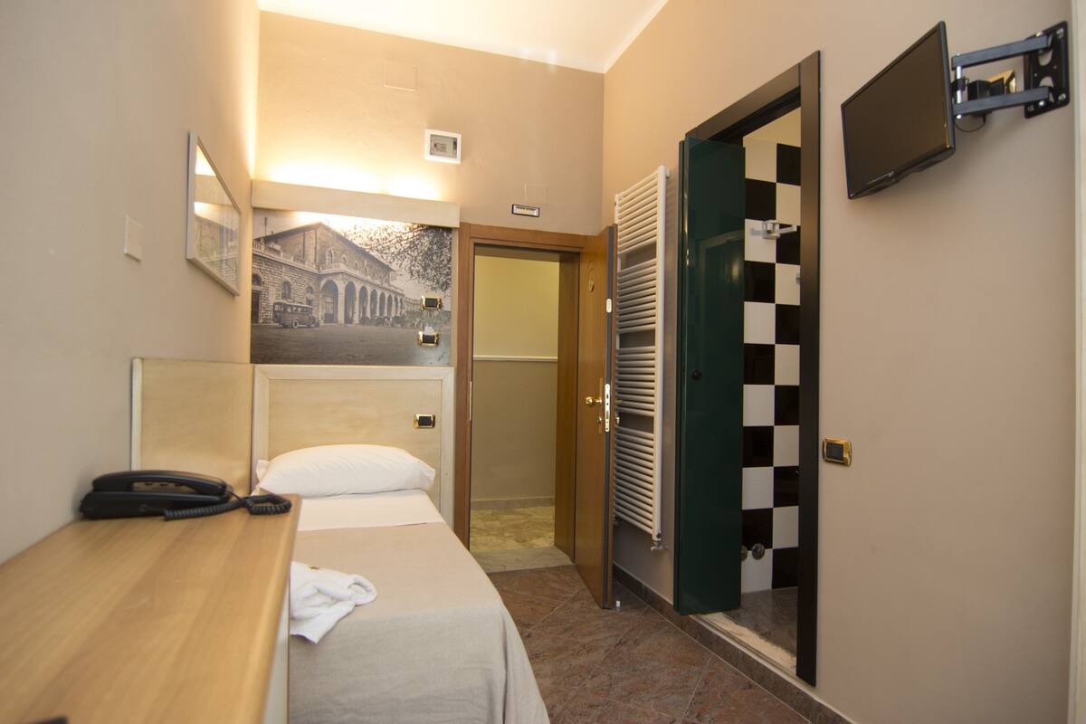 A hotel room with a bed, a bedside telephone, a wall-mounted television, and a door with a checkered pattern.