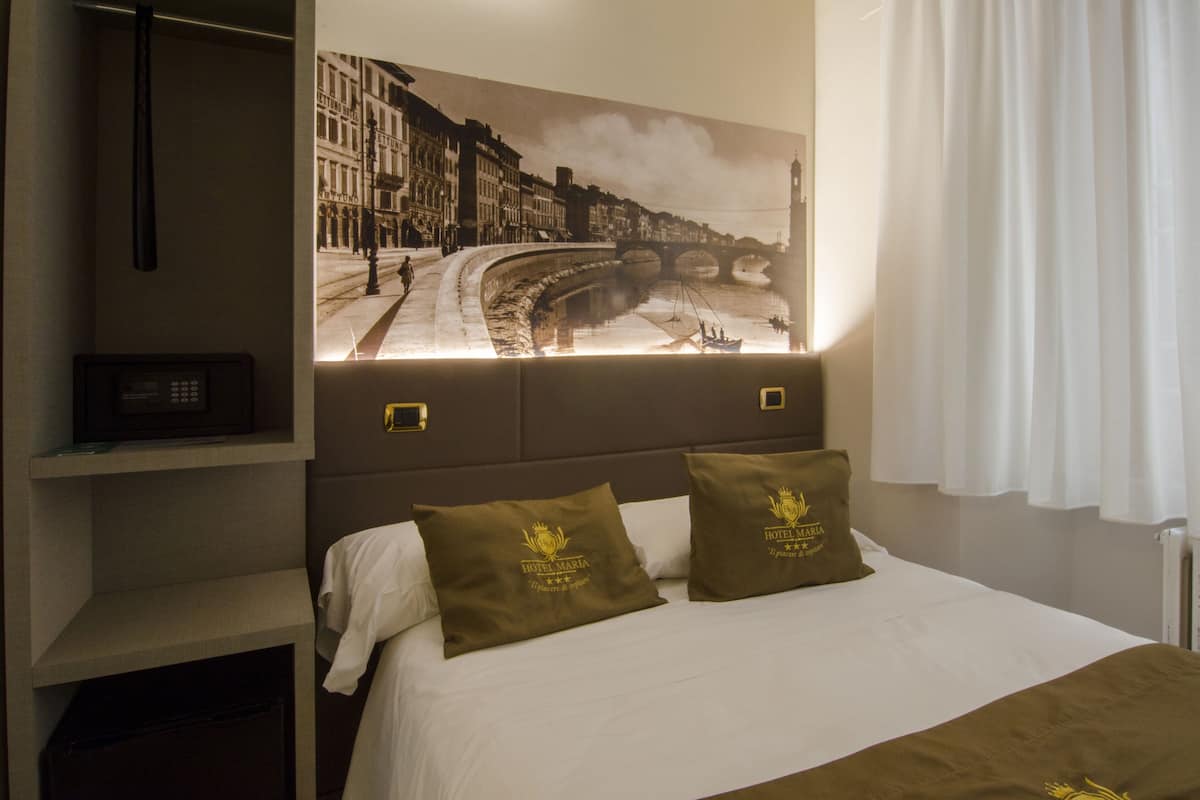 A hotel room with a bed, two pillows, a nightstand, a radio, and a wall-mounted picture of a cityscape.