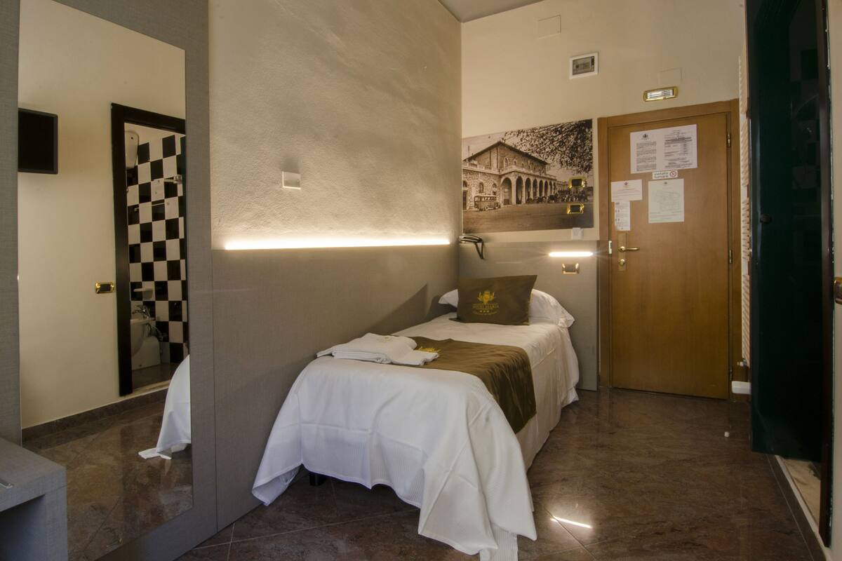 A hotel room with a bed, a checkered mirror, a wall-mounted light, a door with a nameplate, and a poster on the wall.