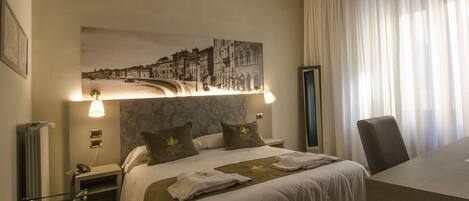 A hotel room with a bed, a desk, a chair, a radiator, a wall-mounted lamp, and a large framed picture.
