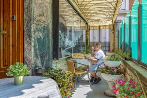 A man sitting at a table outside a building with a large window displaying a cityscape. There are potted plants and a table with a basket of food items. - Hotel Maria (Pisa)