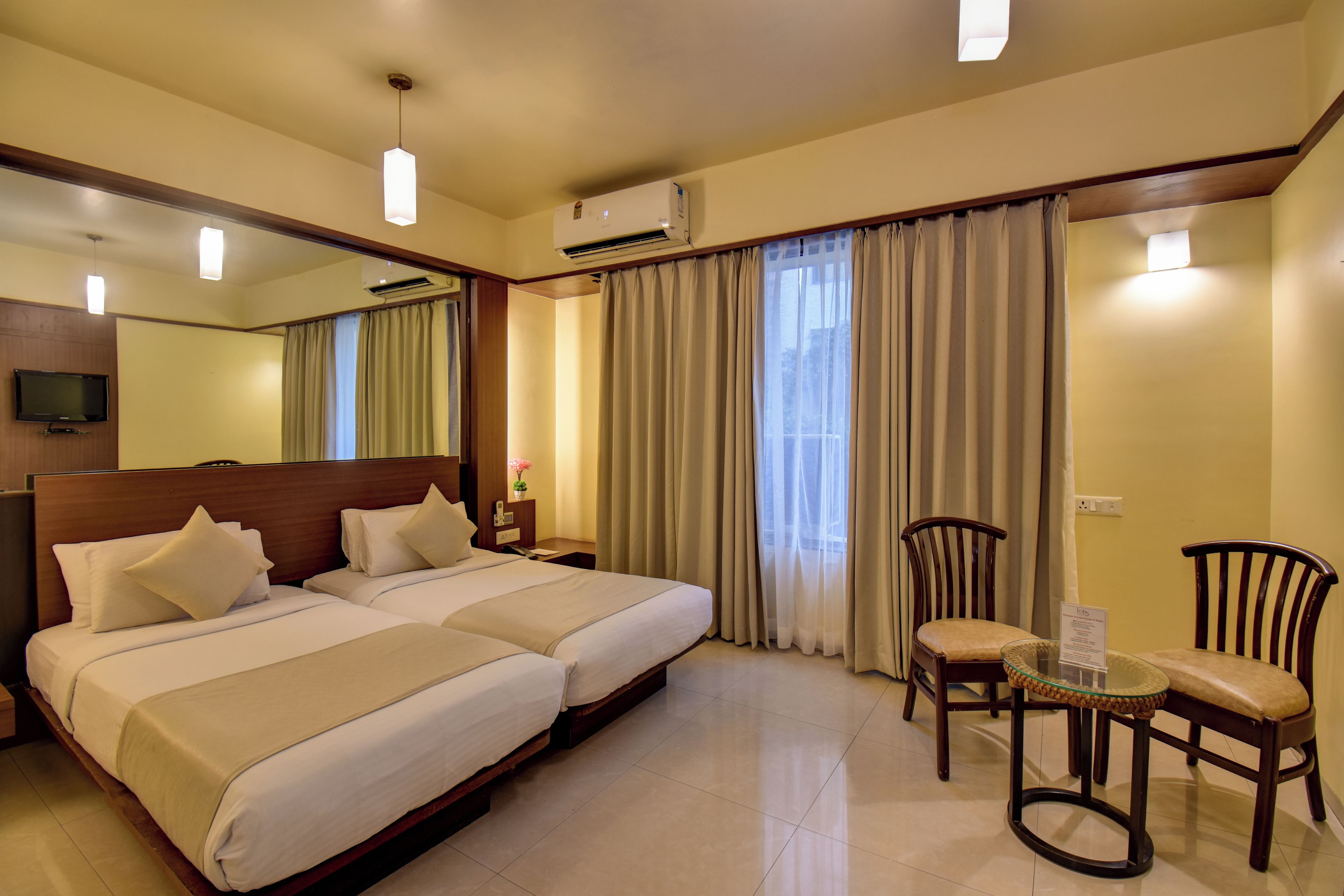 A hotel room with two beds, a TV, an air conditioning unit, a dining table with chairs, and a large window with curtains.