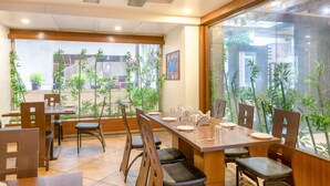 A restaurant with a long wooden table set for dining, surrounded by chairs, and large windows with bamboo plants. - Ivy Studios Hotel Near Pune Airport (Pune)