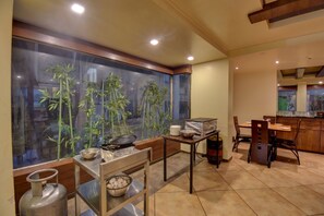 A kitchen with a large window, a gas cylinder, a table with a bowl, and a dining area with chairs and a table. - Hotel Lotus Ivy Studios,Near Pune Airport (Pune)