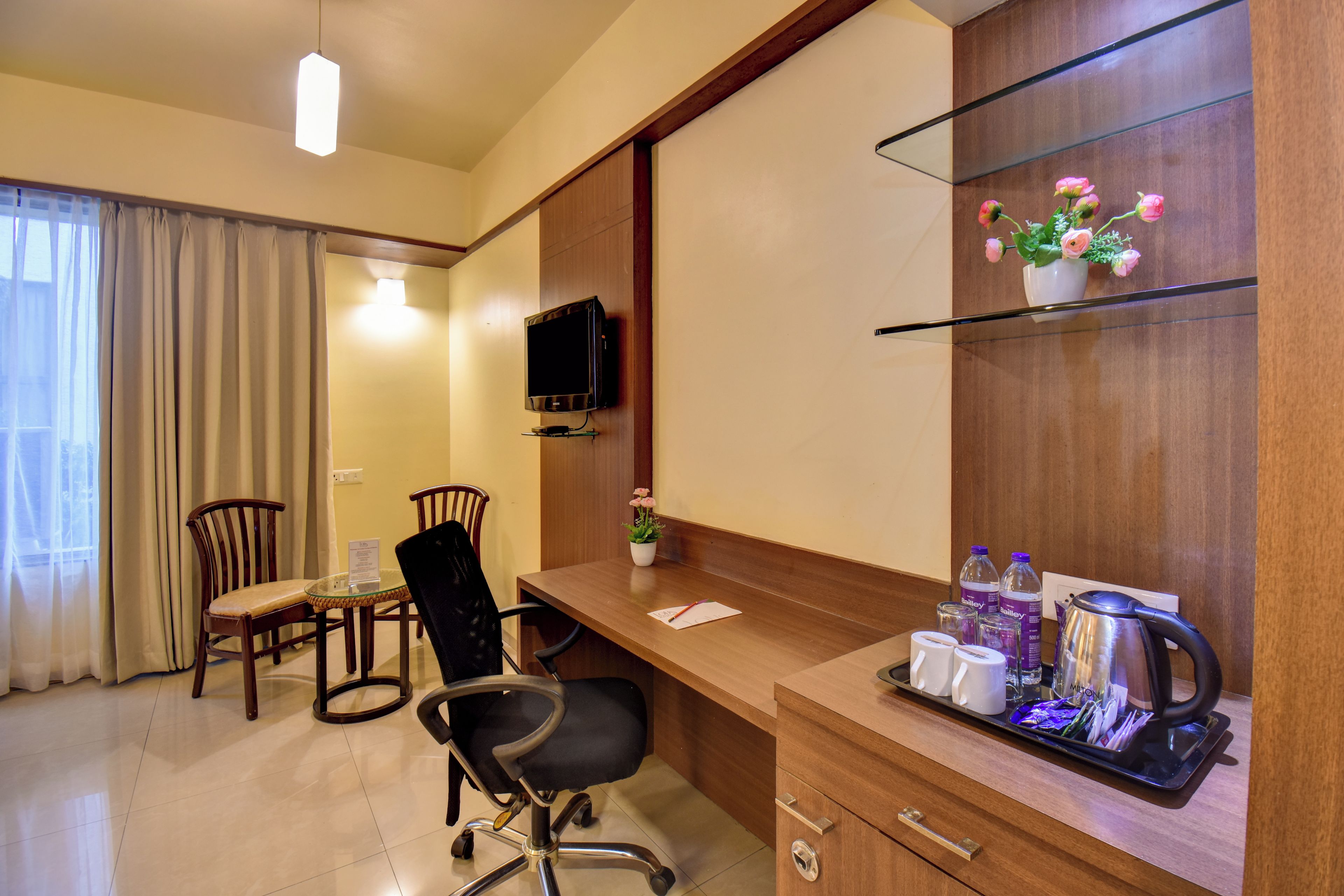 Een hotelkamer met een houten bureau, twee stoelen, een televisie, een kleine tafel met een bloevas, en een dienblad met kopjes en een waterkoker.