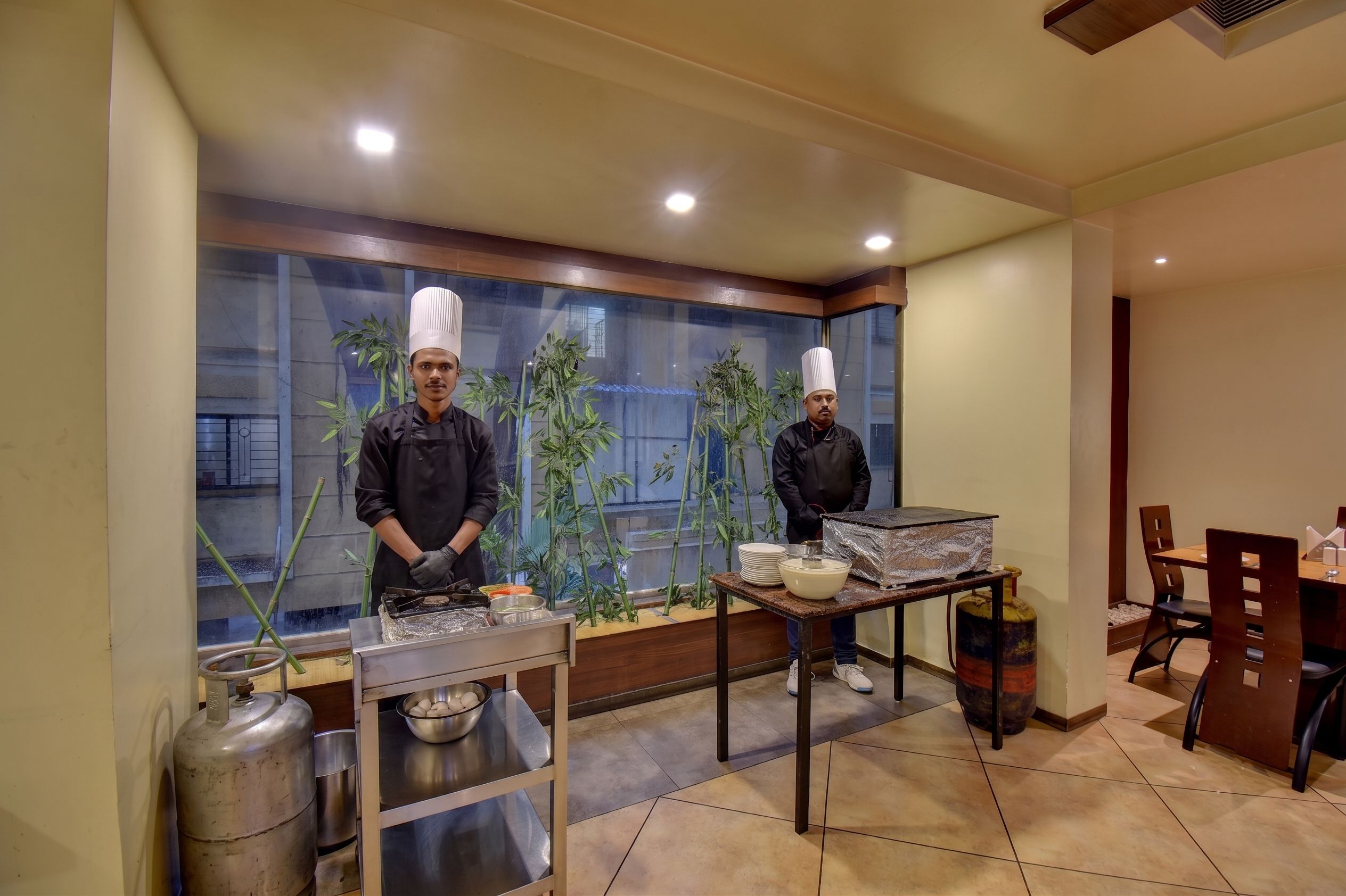 A kitchen with two chefs, a table with cooking equipment, and a dining area with chairs and a table.