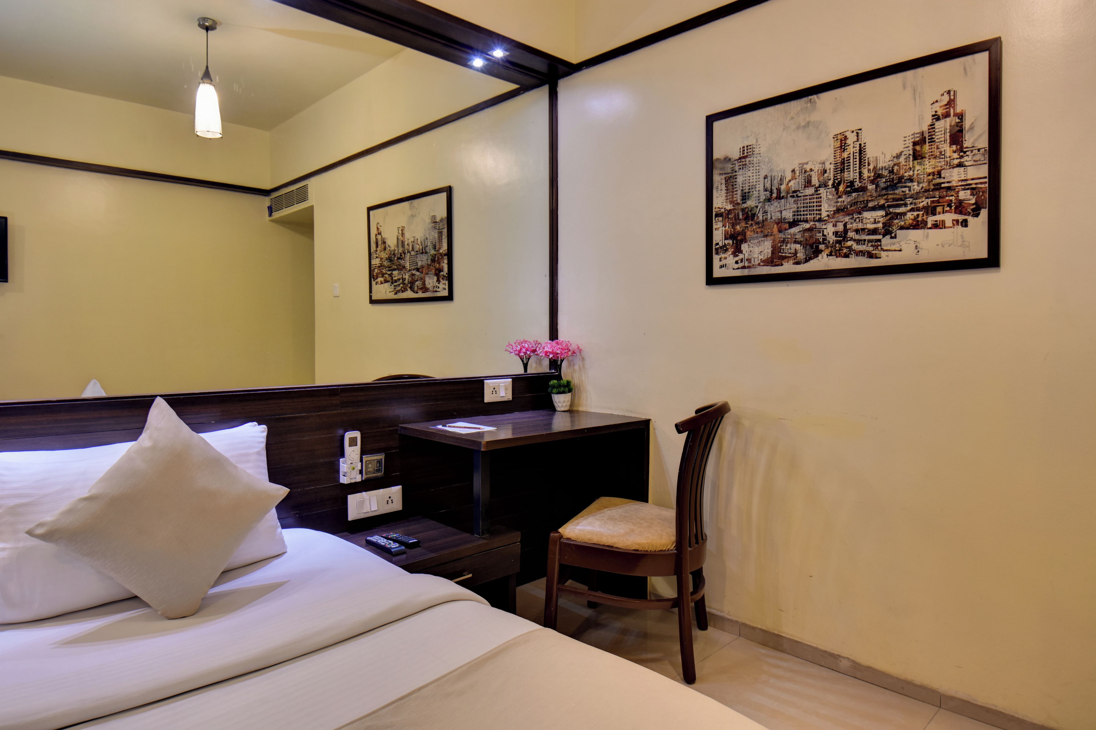 A hotel room with a bed, bedside table, chair, artwork, and a desk with a plant.