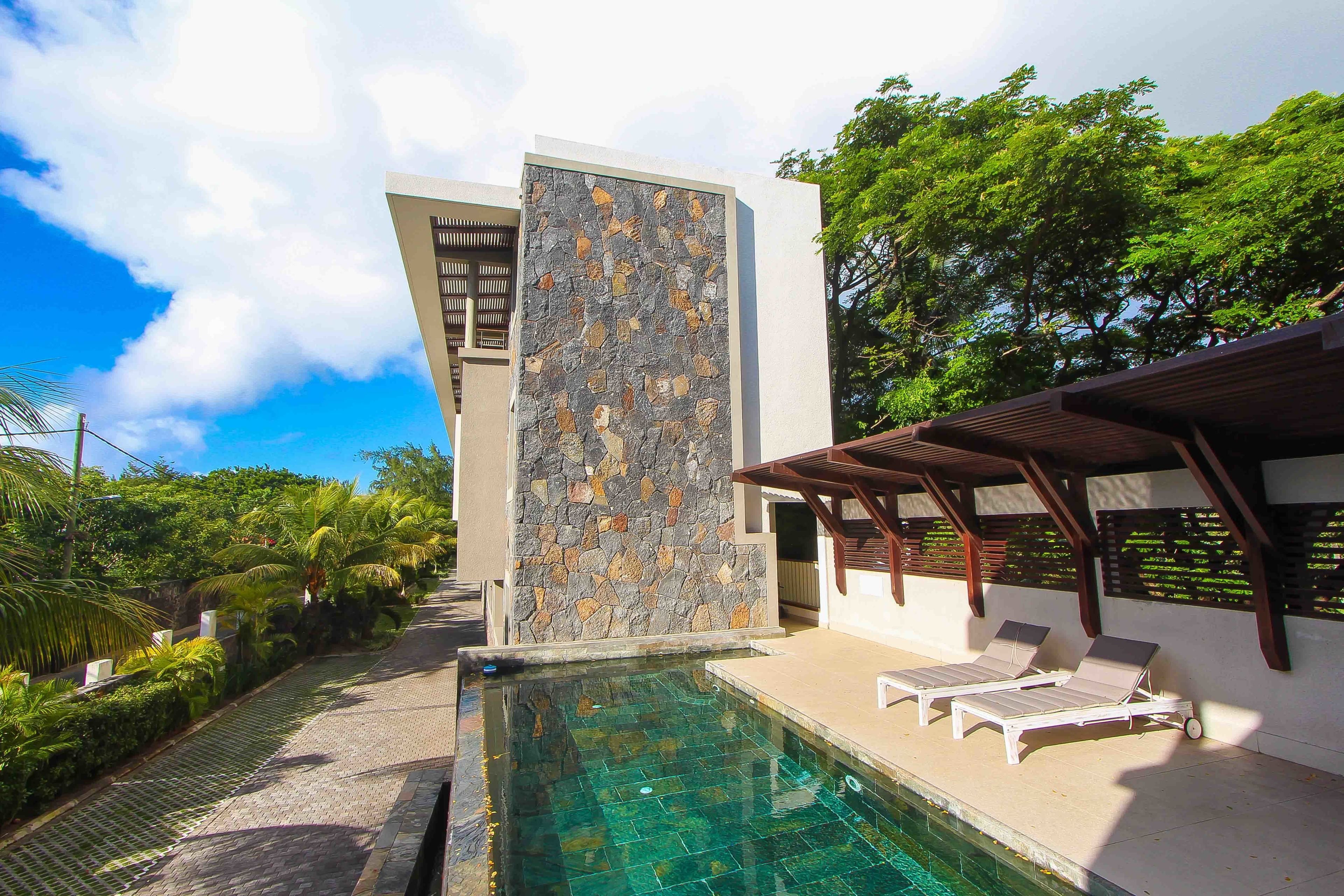 Un área de piscina moderna con un muro de piedra, dos sillones de relax y una pérgola.