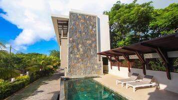 Un área de piscina moderna con un muro de piedra, dos sillones de relax y una pérgola.