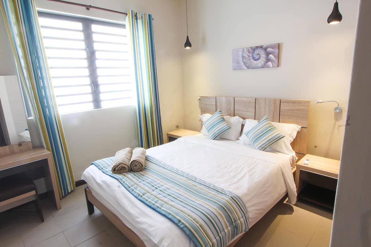 A neatly made bed with striped bedding, a wooden desk with a chair, a bedside table with a lamp, and a window with curtains.