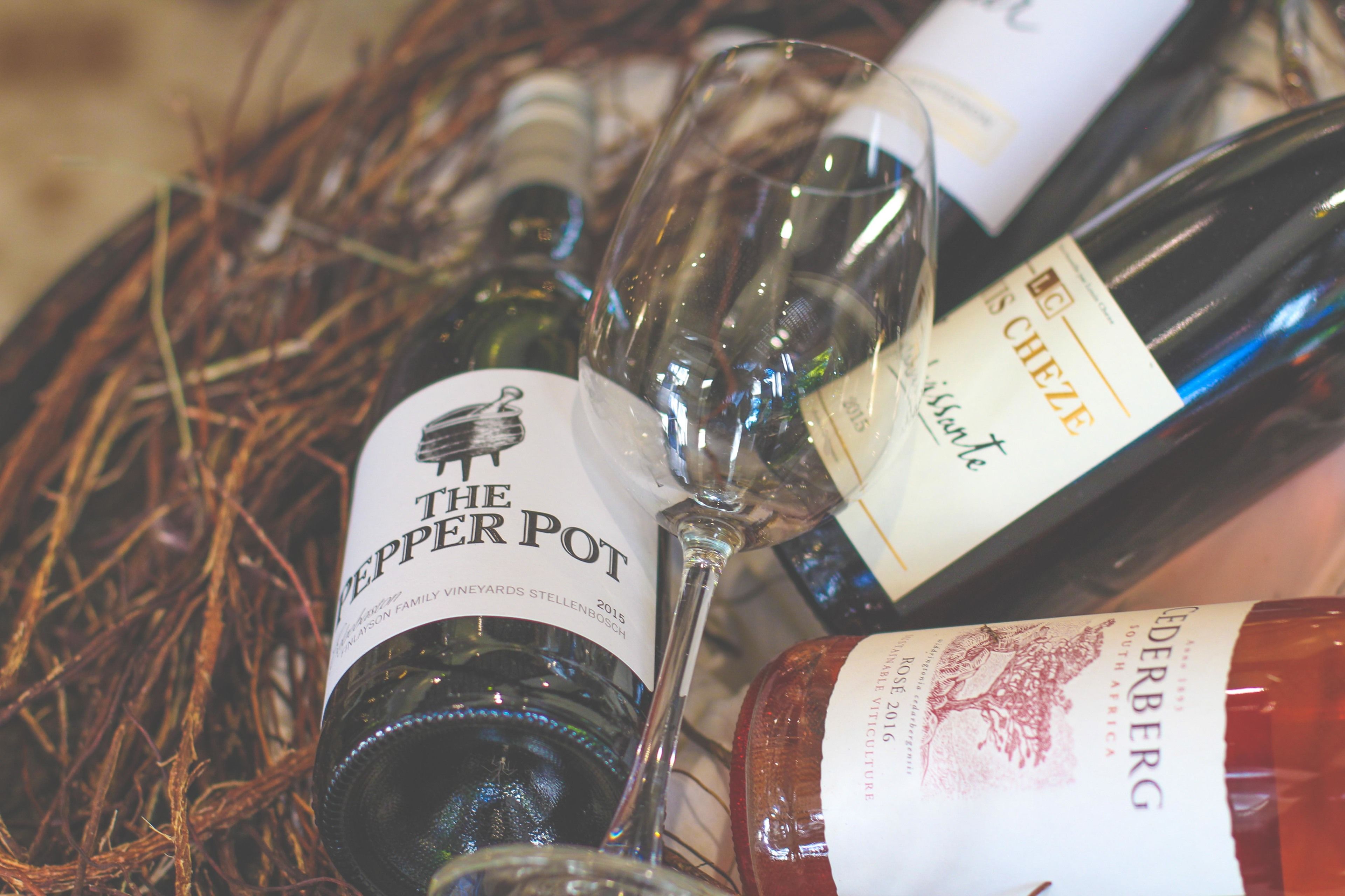 A collection of wine bottles and glasses, including 'The Pepper Pot' and 'Derberg' brands, arranged on a bed of straw.