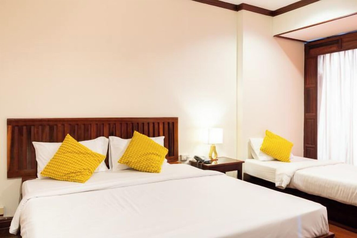 A hotel room with two beds, each with white bedding and yellow pillows. There is a wooden headboard, a bedside table with a lamp, and a window with curtains.