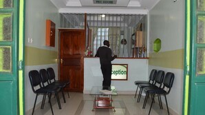 A reception area with a reception desk, two black chairs, a wooden door, and a sign that reads "Reception". - PEC Guest House (Ngong)