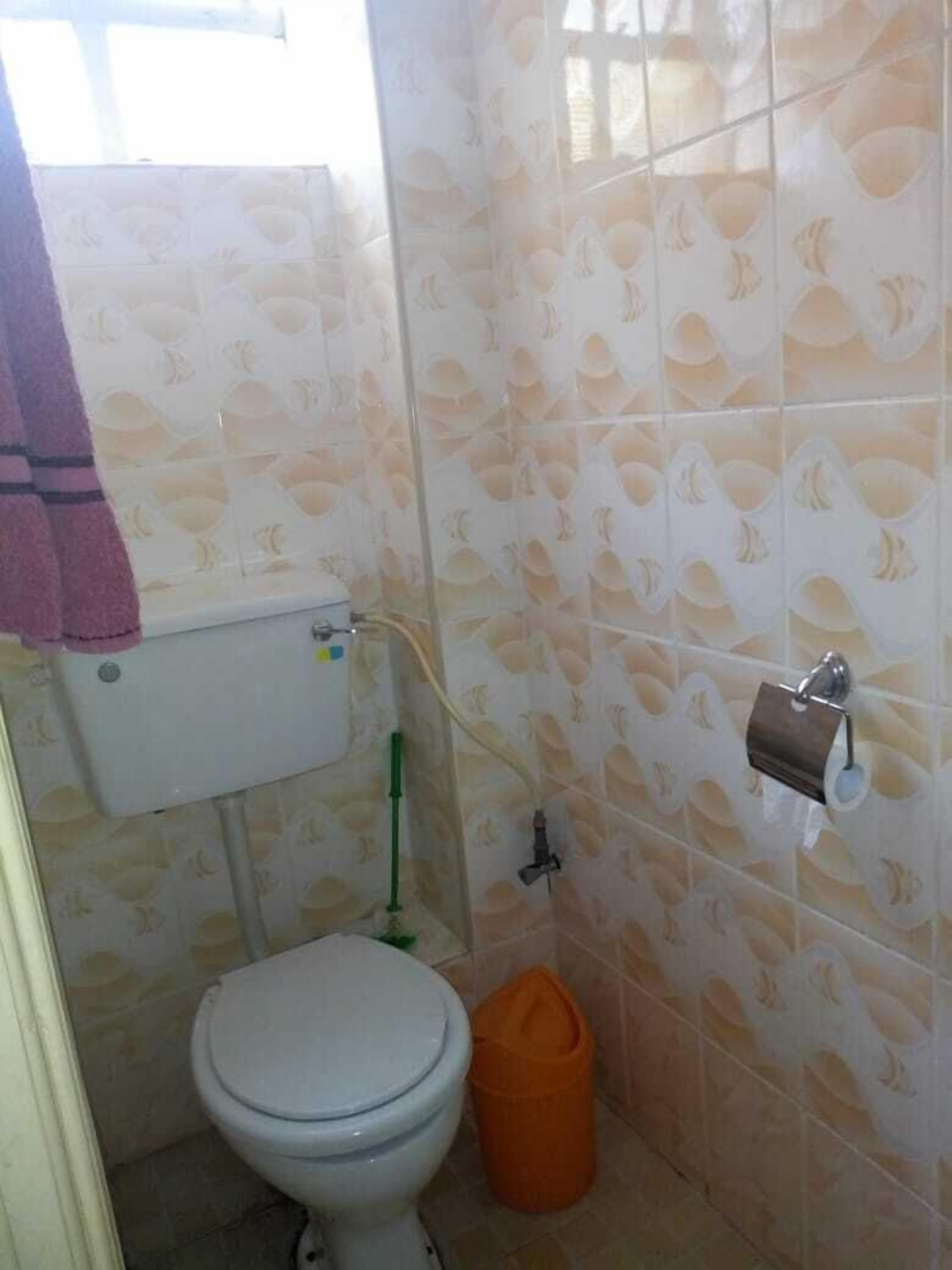 Un baño pequeño con un inodoro, una tubería verde, un rollo de papel higiénico y un cubo de basura naranja.