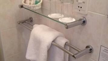 Un baño con un estante de vidrio que contiene artículos de aseo, un perchero con una toalla blanca y un enchufe en la pared.