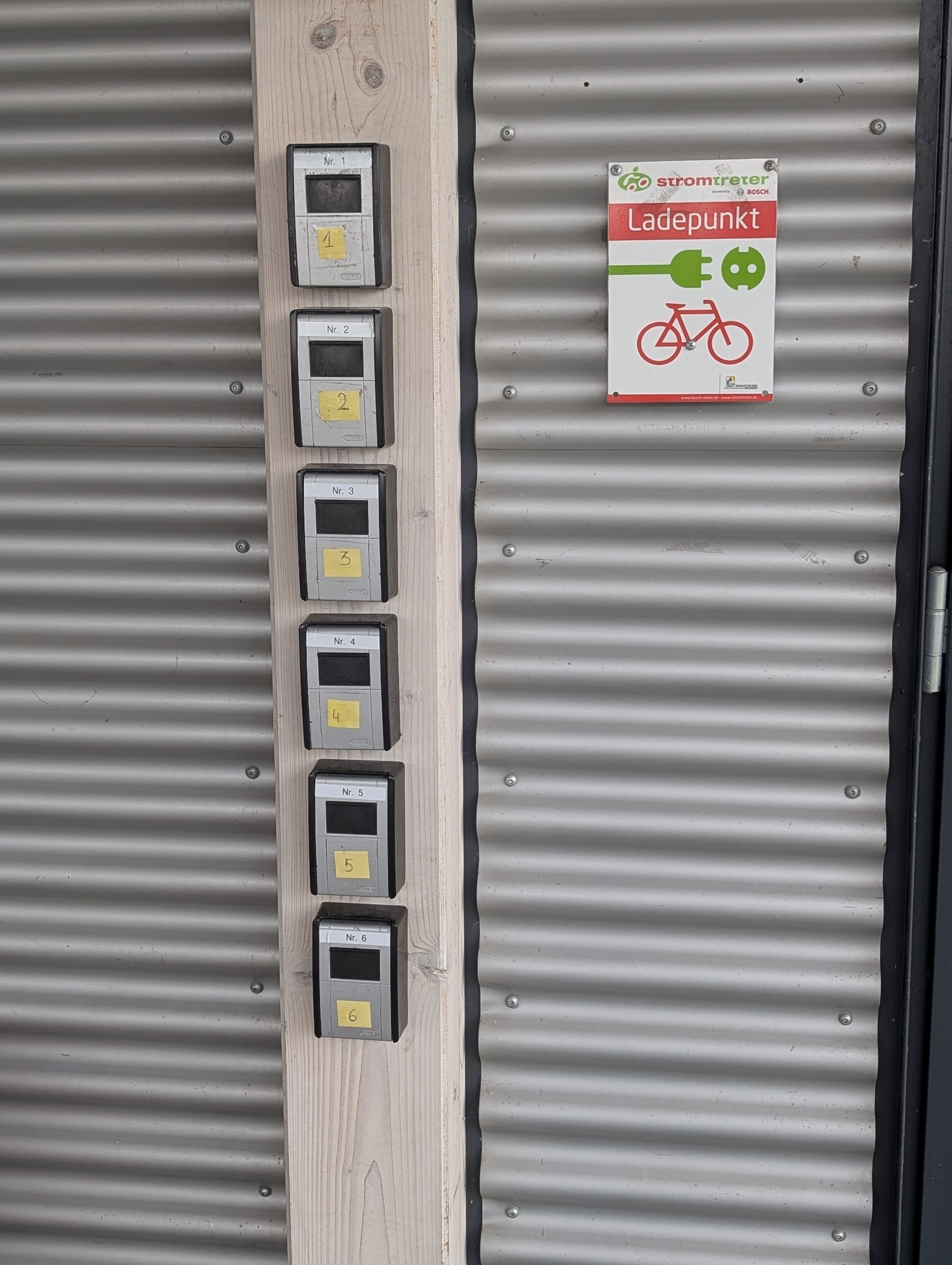 A set of numbered electrical boxes mounted on a corrugated metal wall, with a sign indicating a bike parking area.