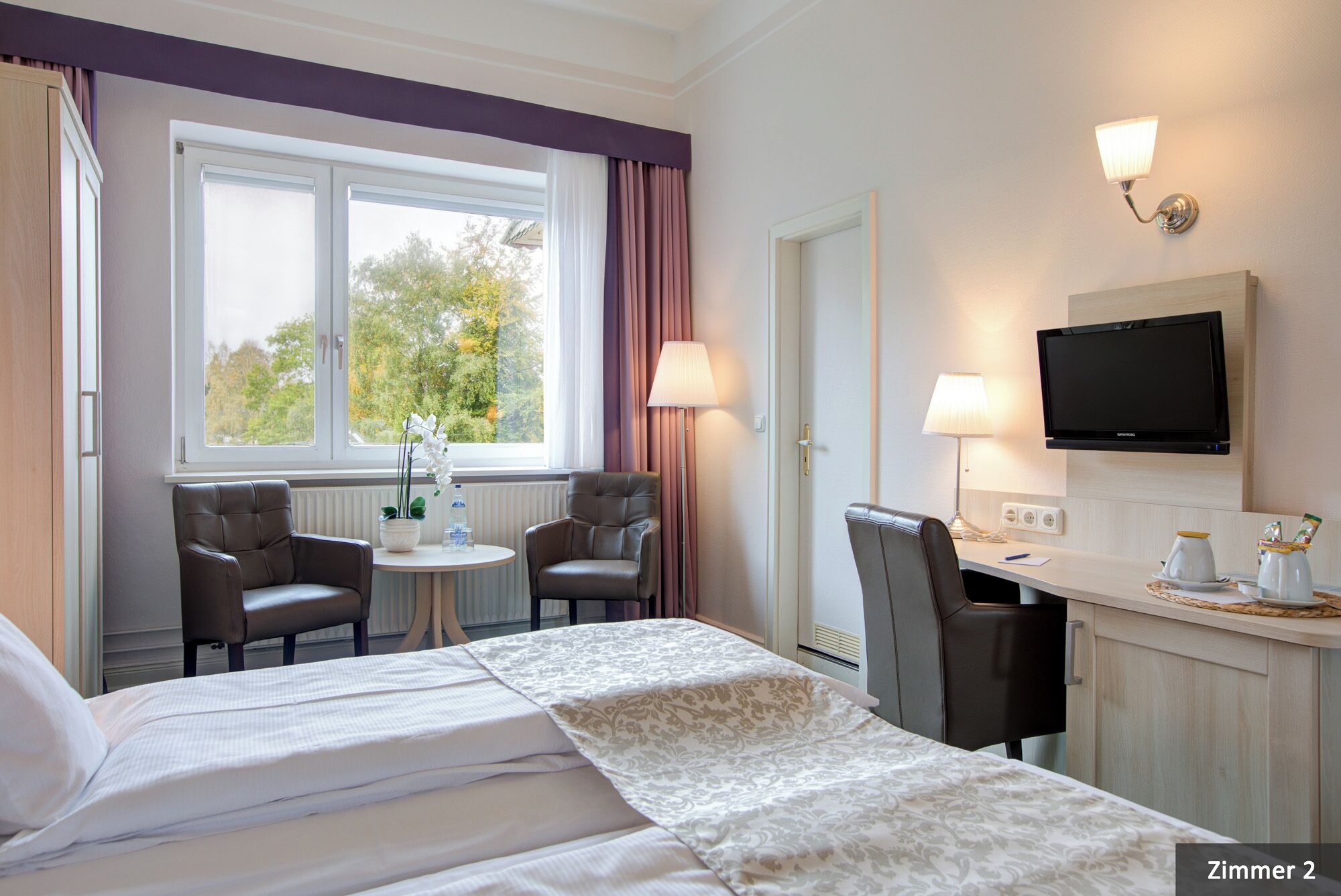 Habitación de hotel con cama, dos sillas, una mesita, televisor y ventana con cortinas.