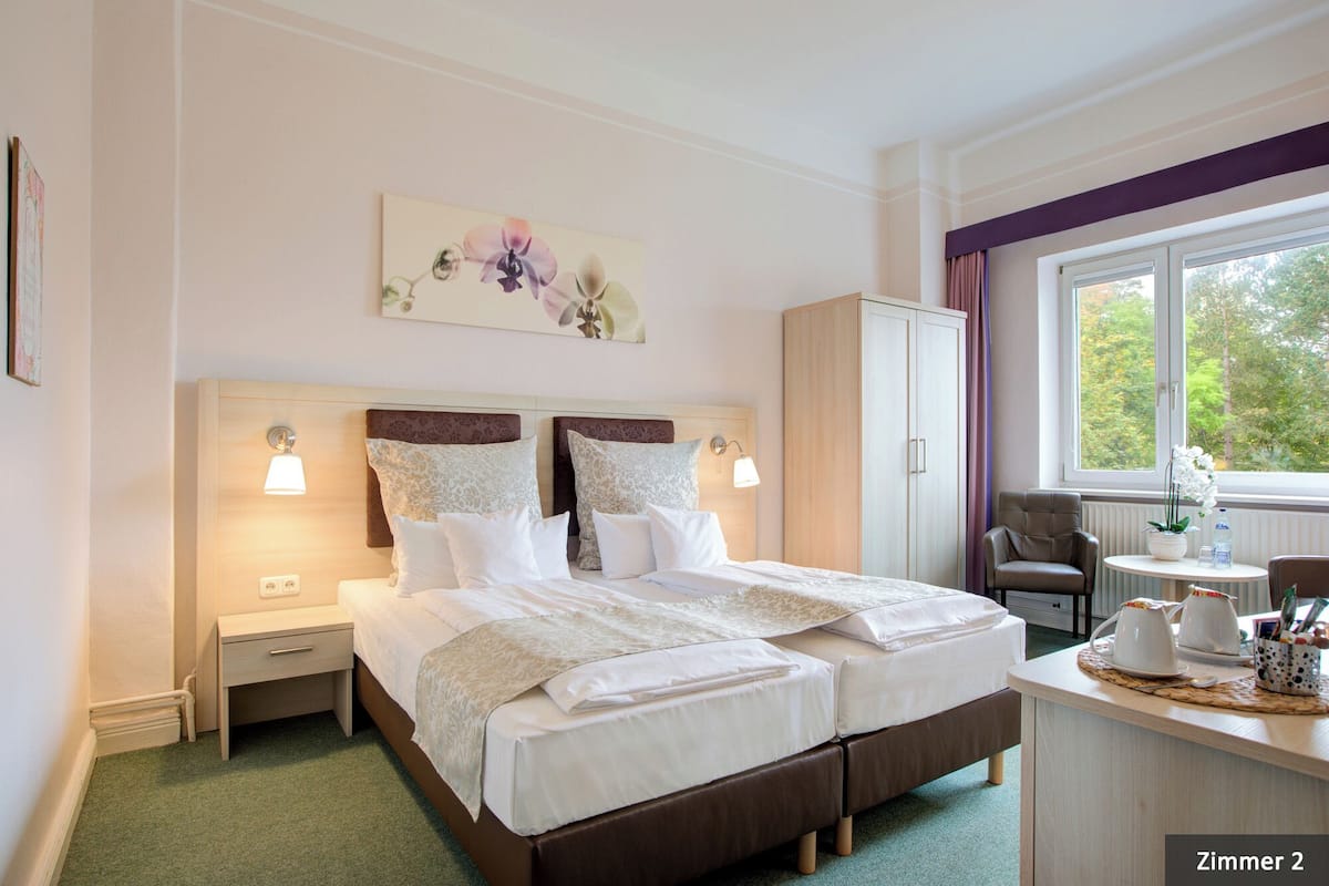 A hotel room with a double bed, bedside lamps, a desk with a chair, a window with a view of trees, and a cabinet.