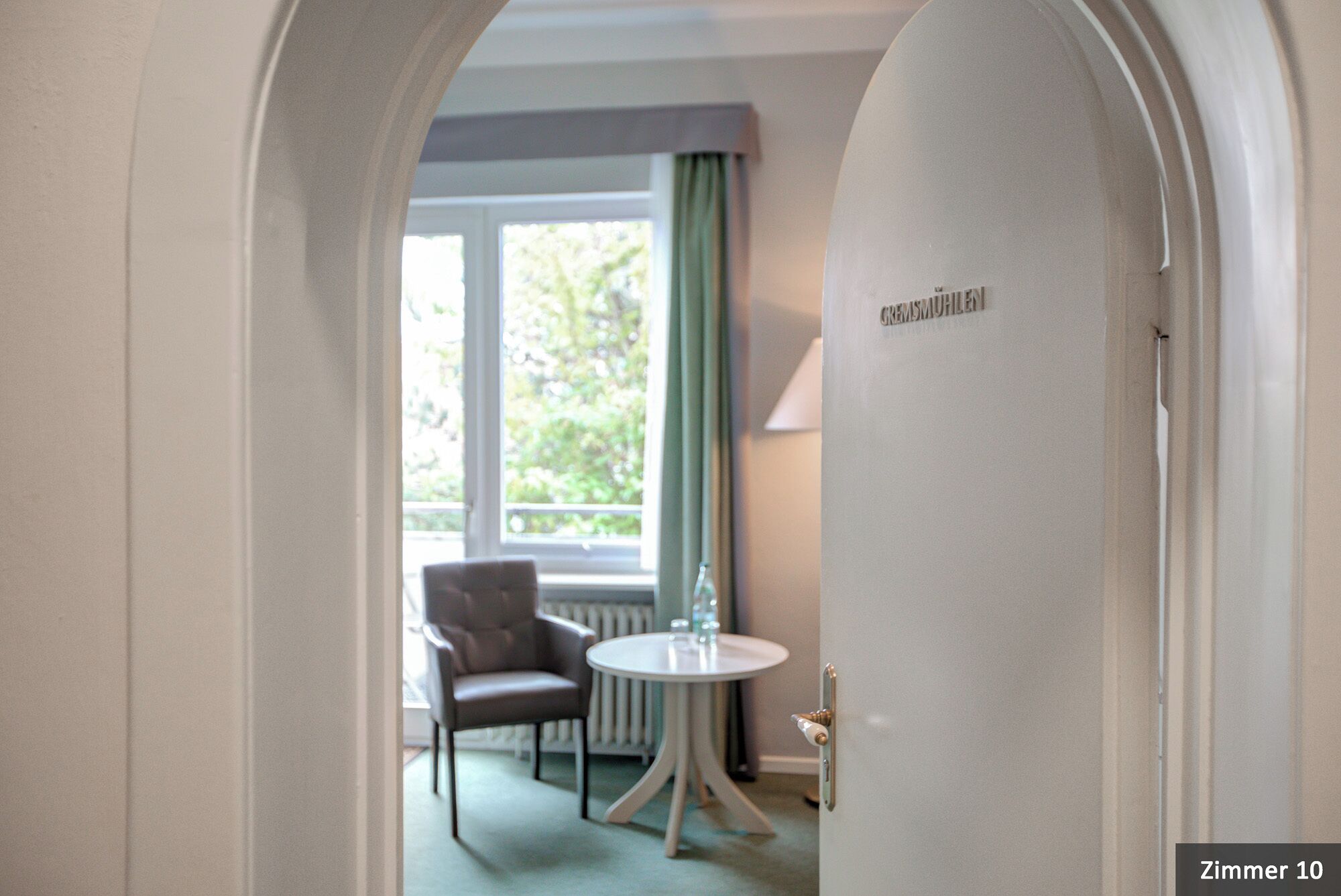 Ein Hotelzimmer mit rundem Tisch, einem Stuhl, einem Heizkörper und einem Fenster mit Vorhängen.
