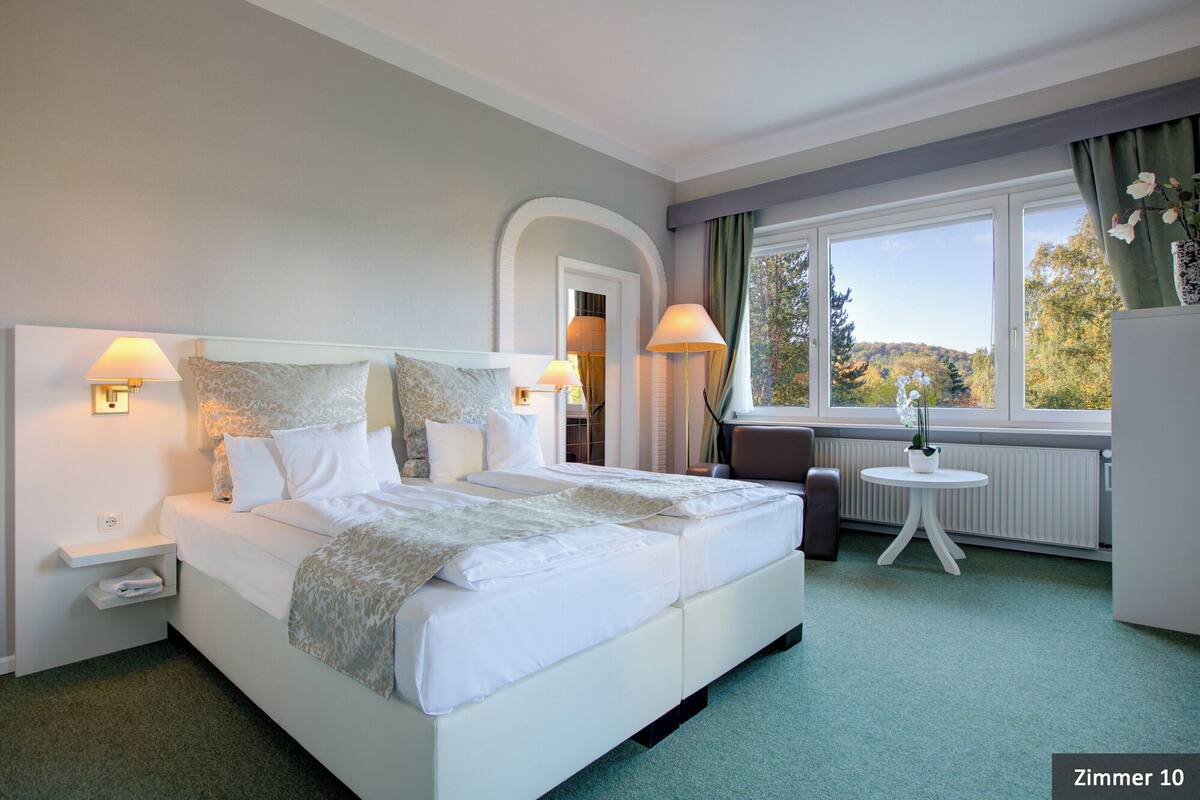 A hotel room with a large bed, two bedside lamps, a small table with a vase, and a view of trees through the window.