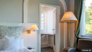 Habitación de hotel con cama, mesita de noche, lámpara, baño con espejo y vistas al exterior.