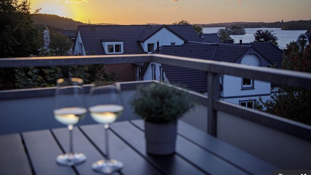 Una terraza con tres copas de vino, una planta en maceta y vistas a casas y un cuerpo de agua al atardecer.