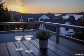 A terrace with three glasses of wine, a potted plant, and a view of houses and a body of water during sunset.