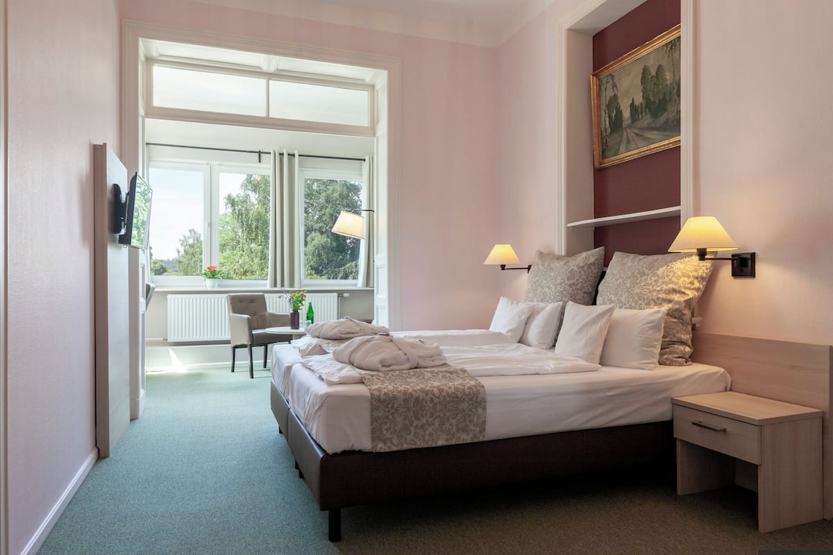 A hotel room with a large bed, bedside tables, a desk, and a window with a view of greenery.