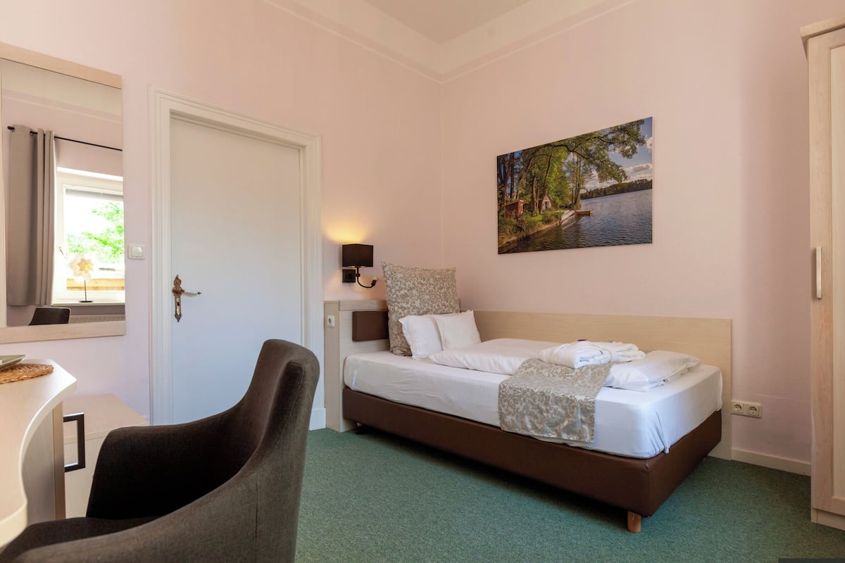 A bedroom with a bed, a desk, a chair, a painting, and a door.