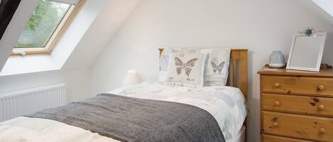 Un dormitorio con techo inclinado, tragaluces, cama con manta gris, almohadas con temática de mariposas, cómoda de madera y mesita de noche con lámpara.