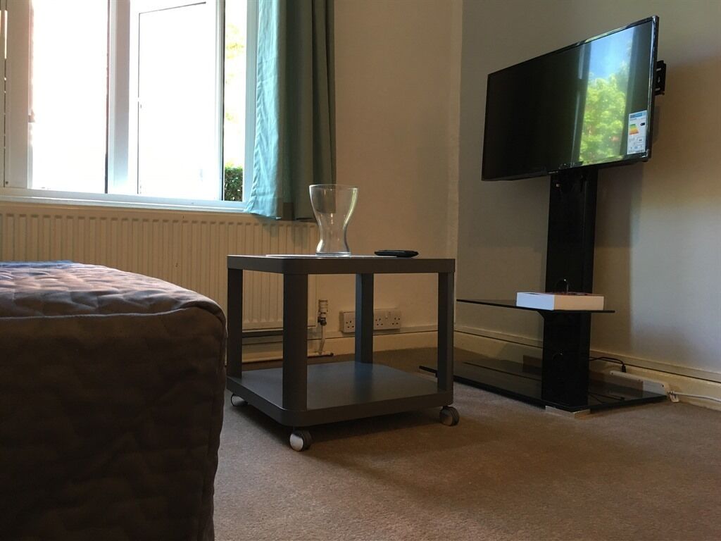 A hotel room with a bed, a small cart with a glass, a flat-screen TV mounted on the wall, and a window with curtains.